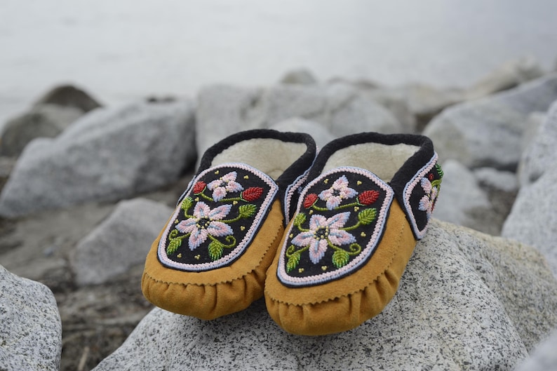 May include: A pair of brown leather moccasins with intricate floral beadwork in shades of pink, white, and green. The moccasins have black trim and are set against a backdrop of gray rocks.