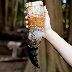 May include: A drinking horn featuring a polished, curved surface with a gradient from light tan to dark brown. The exterior is decorated with intricate white etched patterns, including a central bird motif and geometric designs near the rim.
