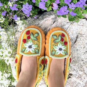 May include: A pair of mustard-colored moccasins with floral beadwork. The design features a large white flower with green and red accents, framed by a green border. The moccasins are displayed outdoors with flowers and rocks in the background.