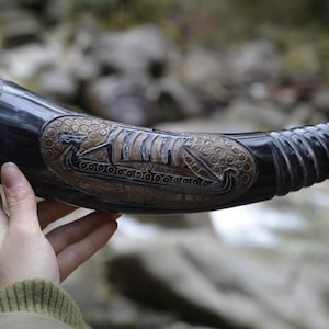 Puede incluir: Un cuerno para beber oscuro y pulido con intrincadas tallas. El cuerno presenta un relieve detallado de un barco vikingo y nudos celtas. El cuerno se sostiene, mostrando su tamaño y artesanía. Aproximadamente 30 cm de largo.