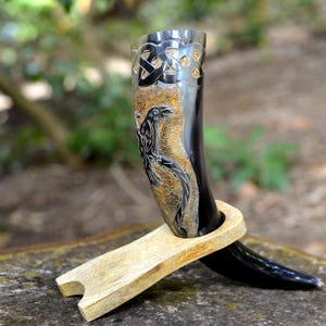 May include: A polished black drinking horn featuring an intricate carved raven design and a Celtic knot pattern near the rim. The horn is supported by a light-colored, smooth wooden stand that holds the curved vessel in an upright position.