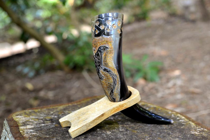 May include: A polished black drinking horn featuring an intricate carved raven design and a Celtic knot pattern near the rim. The horn is supported by a light-colored, smooth wooden stand that holds the curved vessel in an upright position.