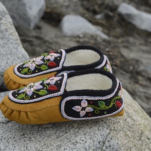 May include: A pair of brown leather moccasins with black and white floral embroidery. The moccasins have a soft, fuzzy lining and are decorated with colorful beads.