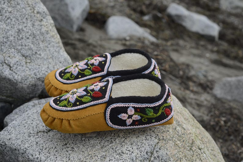 May include: A pair of brown leather moccasins with black and white floral embroidery. The moccasins have a soft, fuzzy lining and are decorated with colorful beads.