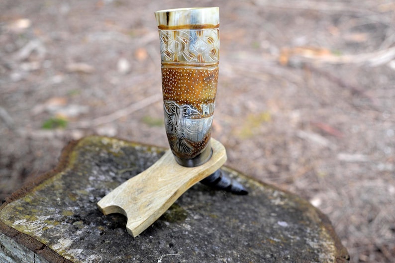 May include: A drinking horn made of polished animal horn with intricate carvings of a bird and geometric patterns, featuring a speckled brown and cream surface, resting on a light-colored wooden stand with a curved cutout design.