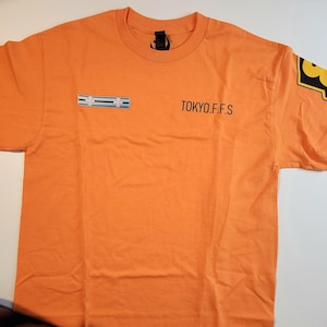 Pode incluir: T-shirt laranja com o texto "TOKYO.F.F.S" em preto. A camisa tem um pequeno design retangular com listras azuis e brancas no peito e um gráfico amarelo e preto na manga. A camisa é feita de um material macio.