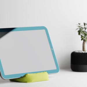 Può includere: Un tablet blu con uno schermo bianco è appoggiato su un supporto verde. Il tablet è su una superficie bianca. Una piccola pianta in vaso è sullo sfondo.
