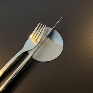 Puede incluir: Un tenedor y un cuchillo de acero inoxidable sobre un plato pequeño y redondo de acero inoxidable. El tenedor está a la izquierda, con el cuchillo en ángulo sobre el plato. El fondo es de color oscuro.