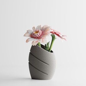 Peut inclure: Un vase gris texturé avec des rainures diagonales contient deux gerberas. Les fleurs ont des pétales roses et blancs et des tiges vertes. Le vase est de conception moderne et minimaliste.