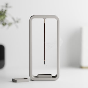 May include: A gray ceramic incense holder with a square base and a rectangular frame. The holder has a small, square opening at the bottom for the incense stick to rest in. The incense stick is suspended in the middle of the frame.