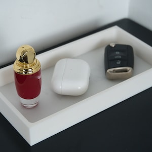 Könnte beinhalten: Ein weißes rechteckiges Tablett mit einer goldenen und roten Parfümflasche, einem weißen kabellosen Ladeetui und einem schwarzen Autoschlüssel mit einem silbernen Knopf.