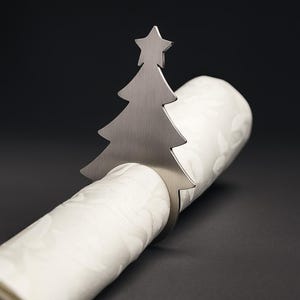 May include: A silver-colored Christmas tree napkin ring. The tree design is cut out and features a star at the top. The napkin ring is wrapped around a white, rolled-up napkin with a subtle floral pattern. The background is dark.