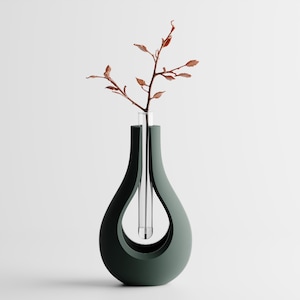 Puede incluir: Un jarrón moderno de color verde oscuro con forma de lágrima, que contiene un tubo de vidrio transparente con una rama delgada de hojas marrones. El jarrón tiene un diseño de corte único, creando un espacio abierto en el centro.