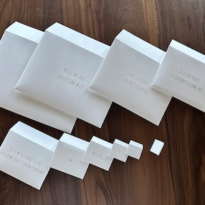 May include: White envelopes with various phrases printed in black, including "Roses are red, violets are blue" and "It is top secret." The envelopes are arranged on a dark wood surface.