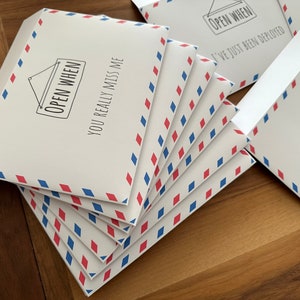 Pu&ograve; includere: Una pila di buste bianche con un motivo a rombi rossi e blu sul bordo. Ogni busta presenta un disegno a linee nere di un cartello appeso con il testo OPEN WHEN e la frase YOU REALLY MISS ME o I'VE JUST BEEN DEPLOYED stampata sotto.