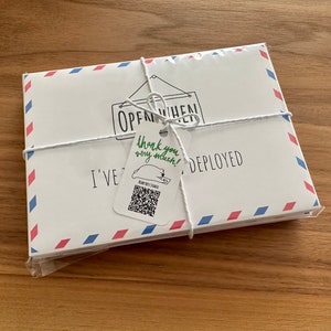 Pu&ograve; includere: Un set di buste rettangolari bianche con bordi in stile posta aerea rossi e blu, legate con uno spago bianco. Un piccolo cartellino attaccato allo spago riporta il testo "thank you very much!" in verde, un codice QR e "I'VE BEEN DEPLOYED".