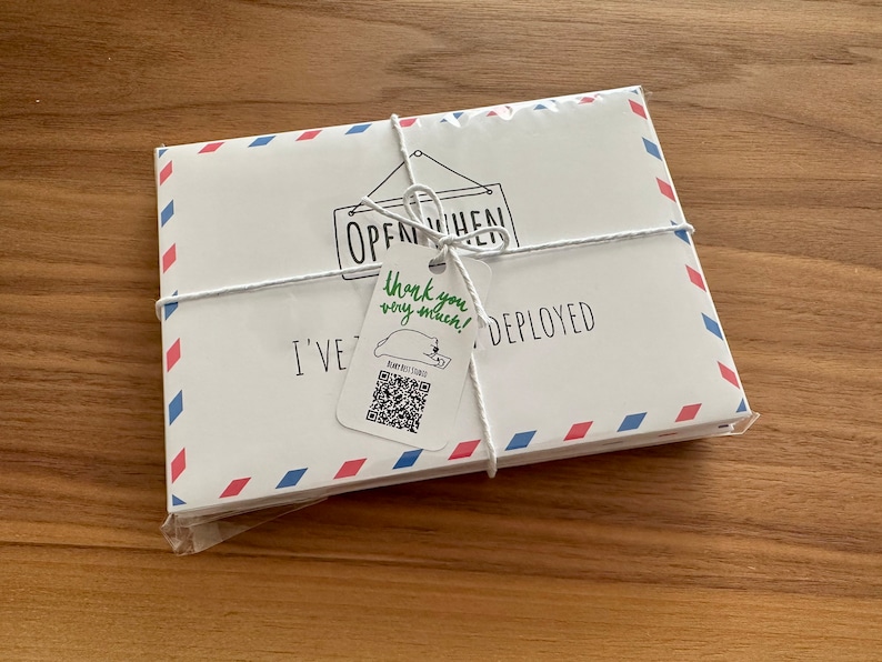 Pu&ograve; includere: Un set di buste rettangolari bianche con bordi in stile posta aerea rossi e blu, legate con uno spago bianco. Un piccolo cartellino attaccato allo spago riporta il testo "thank you very much!" in verde, un codice QR e "I'VE BEEN DEPLOYED".