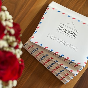 May include: A stack of white envelopes with a red and blue diamond-patterned border. The top envelope features a black line drawing of a hanging sign with the text "OPEN WHEN" and the printed phrase "I'VE JUST BEEN DEPLOYED" below it.