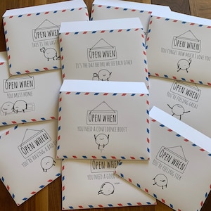 May include: A set of ten white envelopes with red and blue stripes. Each envelope has a handwritten message on the front that says "Open When..." followed by a specific occasion, such as "You Miss Home" or "You're Having a Bad Day".