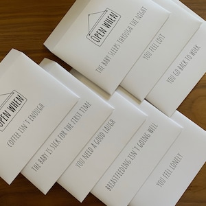 May include: A set of white envelopes with black text. Each envelope features the phrase OPEN WHEN followed by specific scenarios including: The baby sleeps through the night, You feel lost, You go back to work, Coffee isn't enough, The baby is sick for the first time, You need a good laugh, Breastfeeding isn't going well, and You feel lonely.