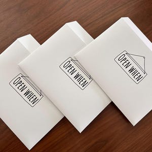 May include: Three white rectangular paper envelopes arranged in a diagonal row. Each envelope features a black printed graphic of a hanging sign with the text OPEN WHEN centered inside the sign, written in a clean, sans-serif black typeface.