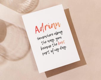 Può includere: Un biglietto di auguri bianco con il nome "Adrian" in scrittura arancione e rossa. Il biglietto include un messaggio scritto a mano con inchiostro nero: "Somewhere along the way, you became the best part of my day."