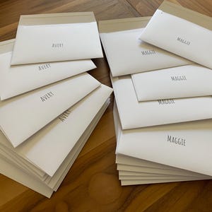 Pu&ograve; includere: Due pile di buste rettangolari in carta bianca. La pila a sinistra presenta il nome AVERY stampato in carattere sans-serif nero su ogni busta. La pila a destra presenta il nome MAGGIE stampato in carattere sans-serif nero su ogni busta.