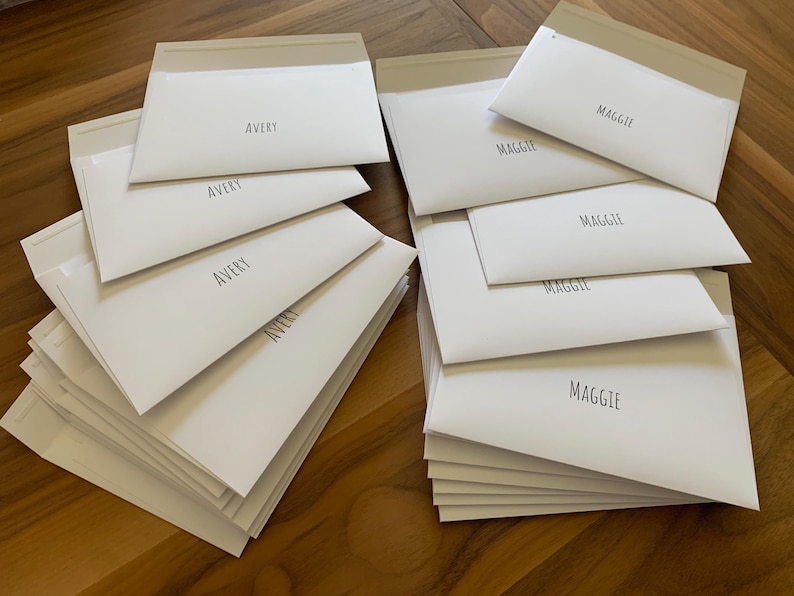 Pu&ograve; includere: Due pile di buste rettangolari in carta bianca. La pila a sinistra presenta il nome AVERY stampato in carattere sans-serif nero su ogni busta. La pila a destra presenta il nome MAGGIE stampato in carattere sans-serif nero su ogni busta.