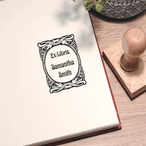 Puede incluir: Un sello Ex-Libris en blanco y negro con el nombre "Samantha Smith" en un marco ovalado decorativo. El sello está sobre una página de libro blanca, con un mango y base de sello de madera visibles.