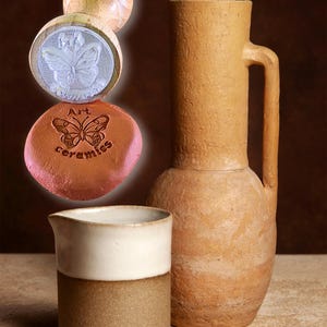 Könnte beinhalten: Eine Sammlung von Keramikartikeln, darunter ein hoher, beigefarbener Krug mit Henkel, ein kleiner weiß-brauner Krug und ein Holzstempel mit einem Schmetterlingsdesign. Der Stempel trägt die Worte "Art ceramics" um den Schmetterling. Die Artikel sind auf einer neutralen Oberfläche angeordnet.