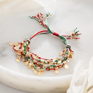 Puede incluir: Un brazalete de múltiples hilos con cuerda roja, blanca y verde y cuentas doradas. El brazalete tiene un borla en cada extremo.