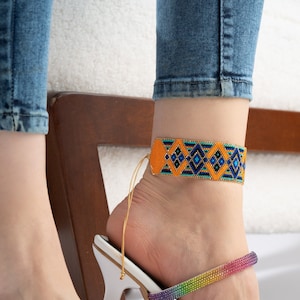 May include: A vibrant orange and blue beaded anklet featuring a geometric design. The anklet is worn on the ankle, and is paired with blue jeans and a white sandal with a rainbow strap.