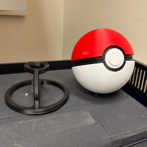 May include: A red, white, and black Poké Ball with a matching black stand. The ball is split into red and white sections with a black band and a white button. The stand is a black, circular base with a central support.