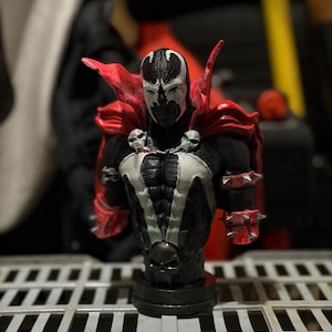 May include: A detailed bust figurine of the character Spawn, featuring a black suit with white chest markings, a flowing red cape, silver spiked wrist gauntlets, and two skull accents on the shoulders, all mounted on a circular black base.