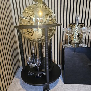 May include: A black drinks trolley with a mirrored disco ball globe, holding champagne bottles and glasses. The trolley has a lower shelf and is on wheels. A champagne bucket and glasses are on a black surface.