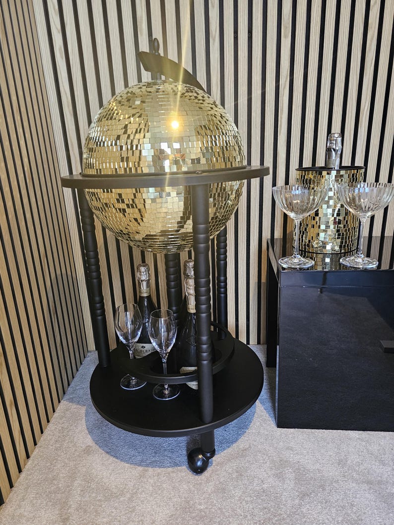 May include: A black drinks trolley with a mirrored disco ball globe, holding champagne bottles and glasses. The trolley has a lower shelf and is on wheels. A champagne bucket and glasses are on a black surface.