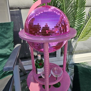 May include: A pink drinks trolley with a mirrored disco ball. The trolley has a round top and bottom shelf, with a bottle of gin and two glasses on the bottom shelf. The cart is outdoors with green plants in the background.