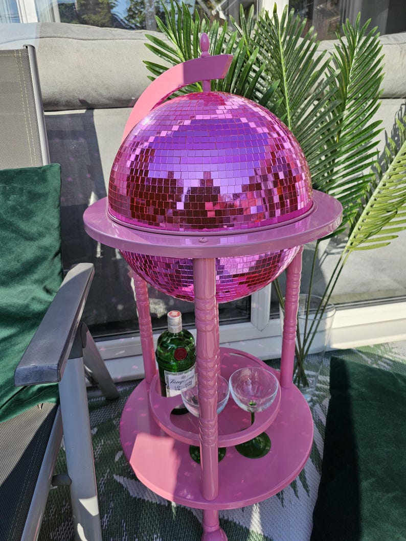 May include: A pink drinks trolley with a mirrored disco ball. The trolley has a round top and bottom shelf, with a bottle of gin and two glasses on the bottom shelf. The cart is outdoors with green plants in the background.