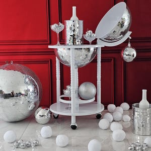 May include: A white drinks trolley with a mirrored disco ball design, holding a bottle of wine, champagne glasses, and a mirrored ice bucket. The trolley is surrounded by white faux snowballs and disco ball baubles, set against a red wall.