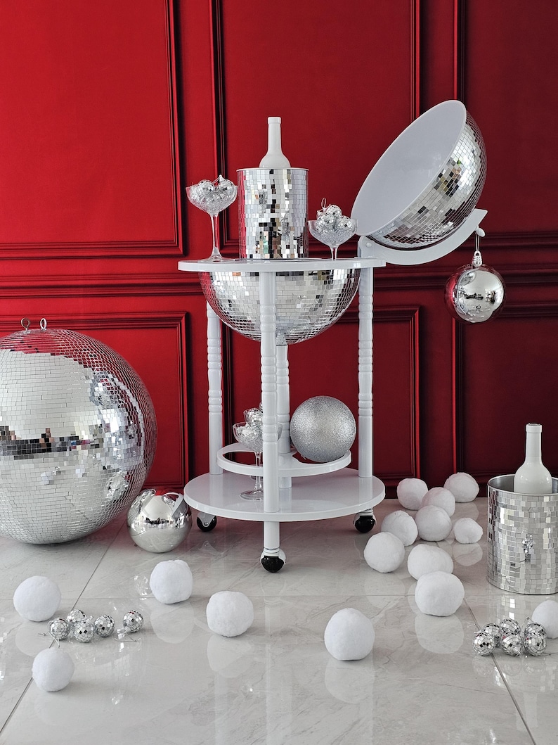 May include: A white drinks trolley with a mirrored disco ball design, holding a bottle of wine, champagne glasses, and a mirrored ice bucket. The trolley is surrounded by white faux snowballs and disco ball baubles, set against a red wall.