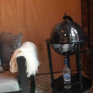 May include: A black drinks cabinet in the shape of a globe, covered in mirrored tiles. A bottle of vodka sits on the lower shelf. A white throw is draped over a black chair. The floor has a geometric pattern.