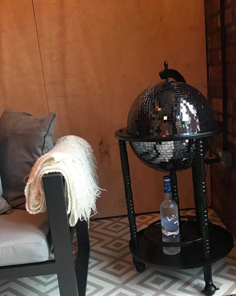 May include: A black drinks cabinet in the shape of a globe, covered in mirrored tiles. A bottle of vodka sits on the lower shelf. A white throw is draped over a black chair. The floor has a geometric pattern.