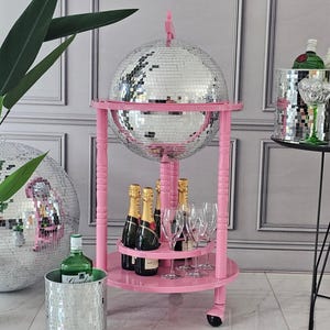 May include: A pink drinks trolley with a large mirrored disco ball, champagne bottles, and glasses. An ice bucket and a green bottle are also present, creating a glamorous party theme.