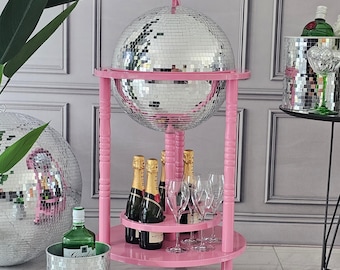 May include: A pink bar cart with a large disco ball on top, holding champagne bottles and glasses. A matching disco ball ice bucket and a green bottle are also visible, creating a glamorous party theme.