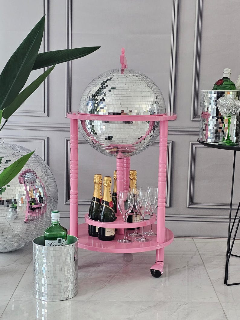 May include: A pink drinks trolley with a large mirrored disco ball, champagne bottles, and glasses. An ice bucket and a green bottle are also present, creating a glamorous party theme.