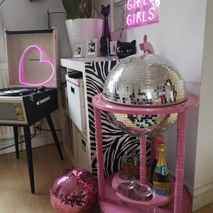May include: A pink drinks trolley with a mirrored disco ball, holding champagne and glasses. A pink disco ball sits on the floor. A record player with a heart-shaped neon light and the text "Girls Girls Girls" in neon are also visible.