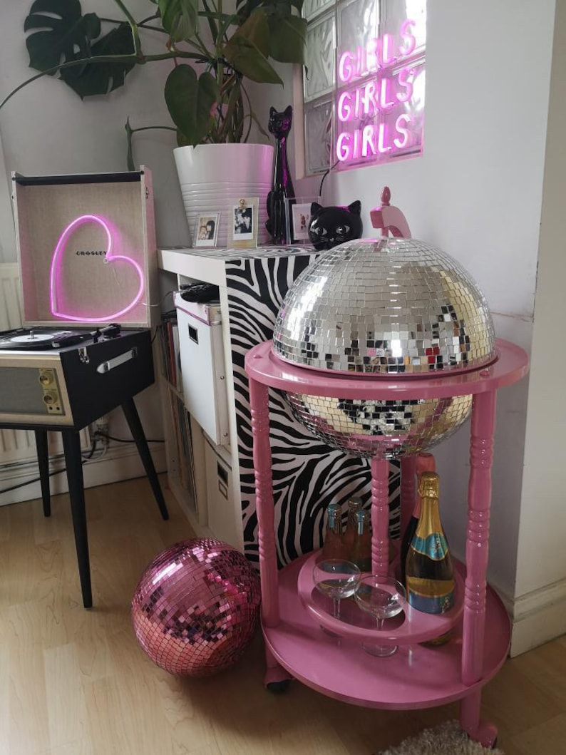 May include: A pink drinks trolley with a mirrored disco ball, holding champagne and glasses. A pink disco ball sits on the floor. A record player with a heart-shaped neon light and the text "Girls Girls Girls" in neon are also visible.