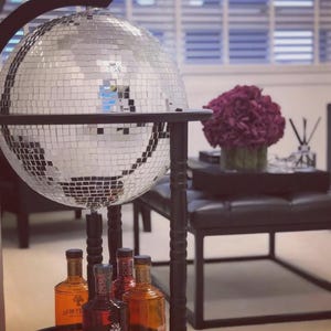 May include: A large, mirrored disco ball hangs from a black stand. Below, several bottles of liquor are displayed on a circular shelf. The bottles contain various coloured liquids and labels. The overall theme is a home bar or entertainment area.