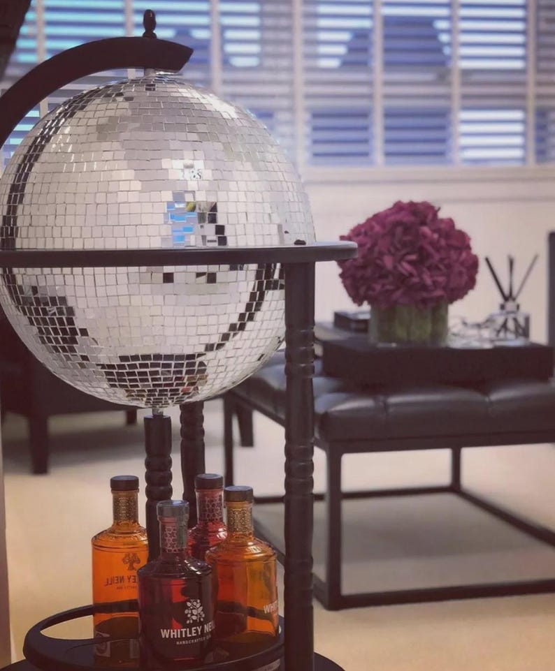 May include: A large, mirrored disco ball hangs from a black stand. Below, several bottles of liquor are displayed on a circular shelf. The bottles contain various coloured liquids and labels. The overall theme is a home bar or entertainment area.