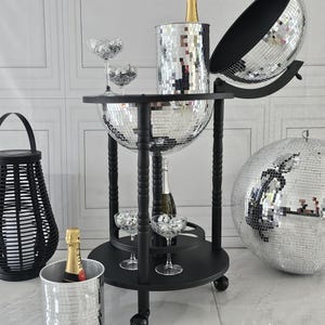 May include: A black drinks trolley with a mirrored disco ball design. It features champagne bottles, glasses, and a disco ball ice bucket. A black woven lantern and a large disco ball are also present.